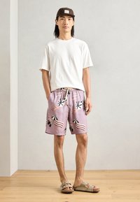 Tenue décontractée composée d'un t-shirt en coton blanc, de shorts de bain lilas avec des motifs de plage et de sandales marron. Design simple, coupe décontractée.
