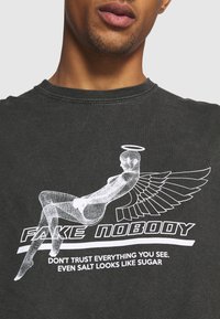 Tmavě šedé tričko s bílým grafickým potiskem postavy s křídly, textem "FAKE NOBODY" a citátem níže. Bavlněný materiál, kulatý výstřih.