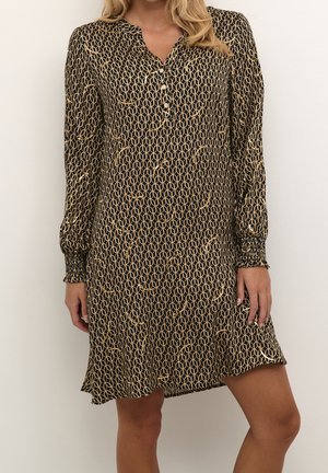 Vestido negro de manga larga con estampado de cadenas doradas, cuello abotonado y puños fruncidos, llevado por una persona con cabello rubio.