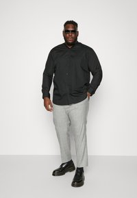 Schwarzes Langarmhemd, graue Karohose, schwarze Schuhe und dunkle Sonnenbrille. Das Outfit hat eine taillierte Passform mit Knopfdetails.