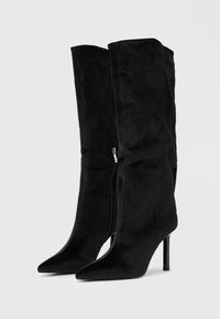 Fekete velúr magas sarkú csizma hegyes orral, térdig érő kialakítással és oldalsó cipzár részlettel. Simított textúrájú, testhez simuló alakzat.