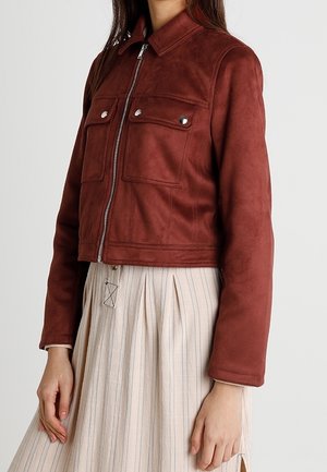 Femme portant une veste courte zippée rouille marron avec deux poches à rabat devant, sur une jupe plissée crème taille haute à rayures verticales.