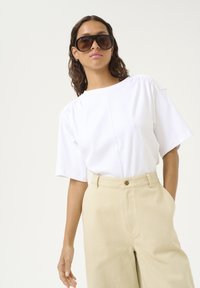 Witte top met korte mouwen en geplooide schouders, gecombineerd met hoog Gesneden beige broek. Model met grote bruine zonnebril.