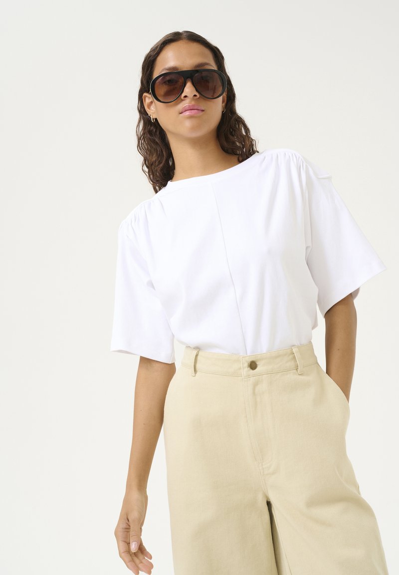 Witte top met korte mouwen en geplooide schouders, gecombineerd met hoog Gesneden beige broek. Model met grote bruine zonnebril.