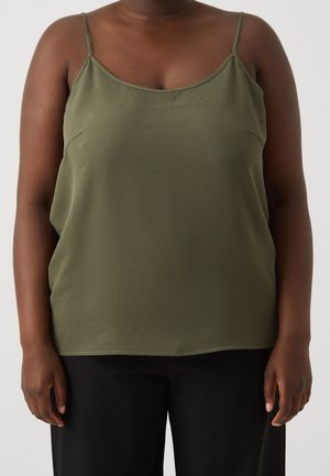 Persoon die een olijfgroene mouwloze top met dunne bandjes en een zwarte broek draagt, staand tegen een eenvoudige lichte achtergrond.