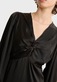 Vestido negro con detalle frontal retorcido, mangas largas con hombros fruncidos y una tela de seda texturizada.