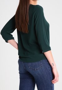 Frau trägt dunkelgrüne Bluse mit dreiviertel Ärmeln und blaue Jeans, steht mit einer Hand in der Hüfte und zeigt ein kleines Handgelenks-Tattoo.