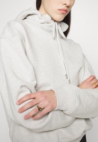 Světle šedá mikina s nařaseným límcem a šňůrkami, se syrovými okraji na rukávech. Ruka drží zlatý prsten ve tvaru tmavé slzy.