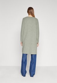 Femme avec de longs cheveux bruns portant un cardigan long gris clair et un jean large bleu, se tenant face à un mur blanc uni.