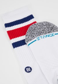 Vita strumpor med röda och blå horisontella ränder; texturerad grå häl med "STANCE" i turkos; cirkulär logotyp på ena sidan.