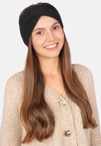 Bandeau en tricot noir avec un design torsadé, fabriqué à partir de matériaux doux, porté par une personne aux longs cheveux bruns, sur un fond blanc.