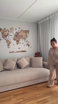 Woman in beige outfit stands beside a beige sofa with pillows, a teddy bear on the armrest, light curtains, and a wooden world map on the wall.