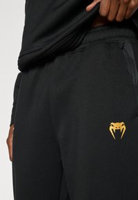 Μαύρο αθλητικό παντελόνι με υφασμάτινη υφή, ελαστική μέση και χρυσό λογότυπο φιδιού στον αριστερό μηρό.
