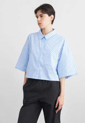 Personne aux cheveux courts et foncés portant une chemise ample à manches courtes à rayures verticales bleu clair et blanc, et un pantalon noir, posant avec une main dans la poche.