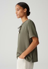 Blusa corta a maniche corte color verde oliva con scollatura rotonda, dettaglio con fiocco, texture liscia e vestibilità rilassata, abbinata a pantaloni chiari.