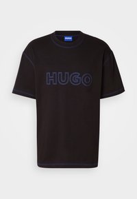 Melna kokvilnas t-kreklā ar lielu, zilu izšūtu "HUGO" logo uz priekšpuses, ar kontrastējošu zilu šuvju apdari.