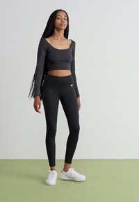 Černý dlouhý rukáv crop top spárovaný s černými legínami, které mají vysoký pas a malé logo. Model má na sobě bílé tenisky na zelené podlaze.