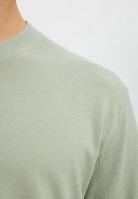 Nahaufnahme eines hellgrünen Rundhalspullovers, getragen von einer Person mit sichtbarem Hals und Schulter vor einfarbigem Hintergrund.