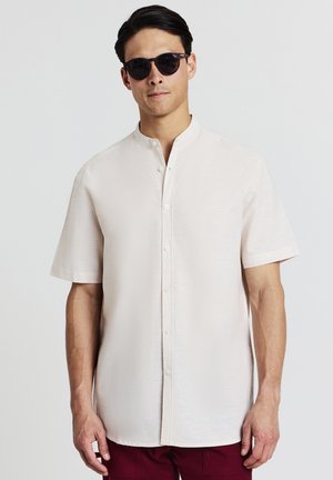 Uomo che indossa una camicia bianca a maniche corte, con bottoni, occhiali da sole scuri e pantaloni rossi scuri su uno sfondo chiaro e semplice.