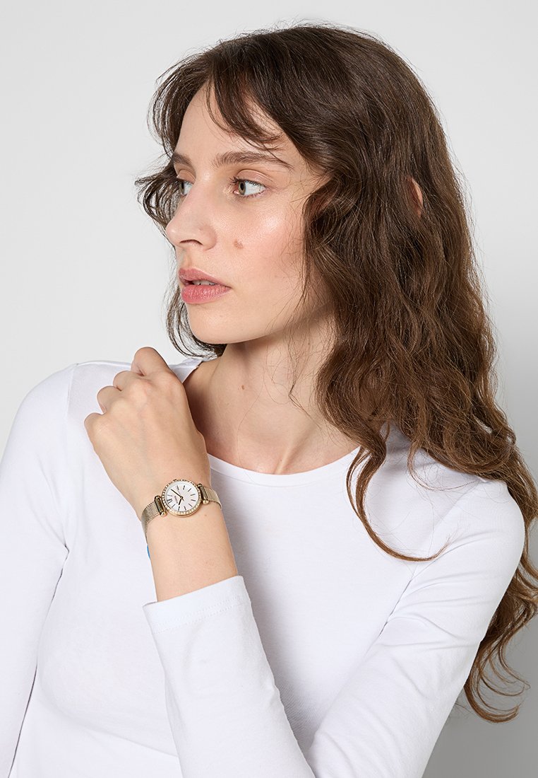 Orologio da donna con cinturino in rete argentata, quadrante rotondo bianco, dettagli dorati e indici neri, indossato su un polso con una maglietta bianca a maniche lunghe.