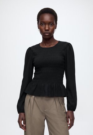 Femme portant un haut noir smocké à manches longues avec un ourlet à basque et un pantalon beige plissé, debout devant un fond clair uni.
