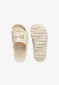 Beige slide sandals made of rubber. Features a textured footbed and a grid-patterned outsole. Gold crocodile logo and brand name embossed.