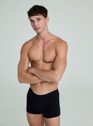 Młody mężczyzna o krótkich brązowych włosach, bez koszulki, w czarnych bokserkach, stojący ze skrzyżowanymi ramionami na jasnym, jednolitym tle.