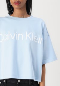 Ljusblå croppad t-shirt med "Calvin Klein" tryckt i fetvita bokstäver. Har en avslappnad passform, korta ärmar och en mjuk textur.