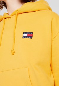 Sudadera con capucha amarilla con cordón y un gran bolsillo. Presenta un pequeño parche de marca con acentos en azul marino, blanco y rojo en el pecho.
