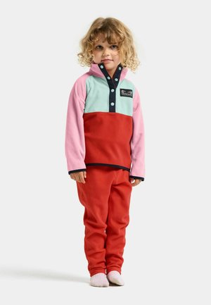 Kind met krullend blond haar draagt een kleurrijk fleecejack met roze, rood en lichtblauwe panelen, bijpassende rode fleecebroek en witte sokken.