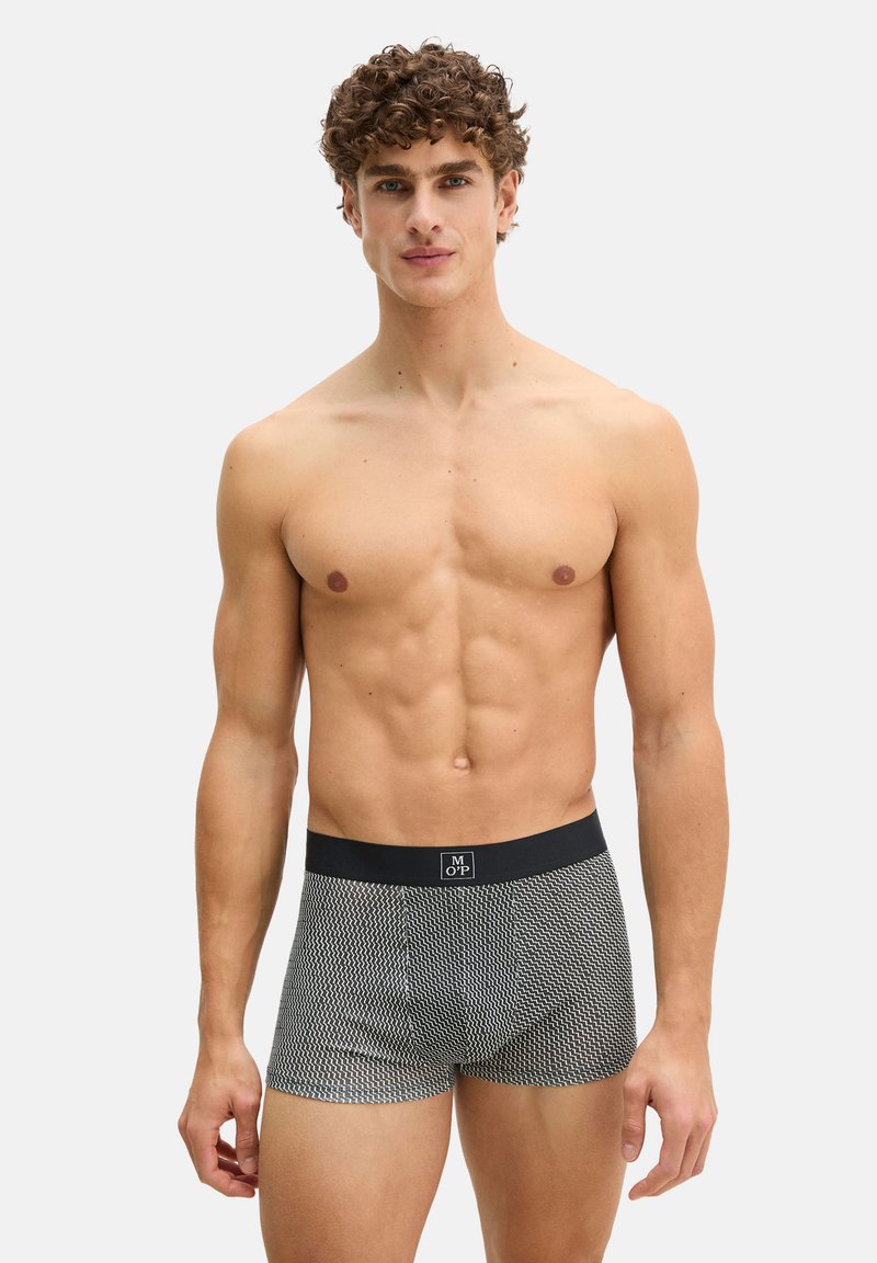Homme musclé aux cheveux bouclés, debout face à l'avant, portant un boxer noir et blanc à motifs avec une ceinture noire.