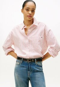 Camisa de botones a rayas rojas y blancas con cuello, mangas enrolladas y un bolsillo en el pecho, combinada con jeans azules y un cinturón negro.