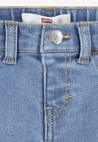 Világoskék farmernadrág sárga varrással, ezüstszínű fémbottonnal a deréknál, és egy fehér címkével a tetején.