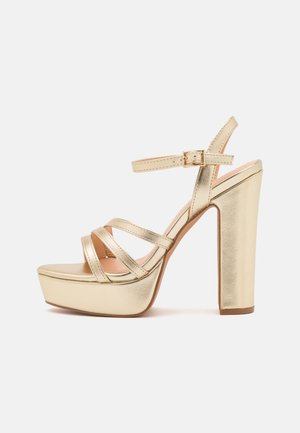 Gold metallic high-heeled sandal with thick platform, adjustable ankle strap, and crisscross straps on the vamp. Smooth texture, structured design.