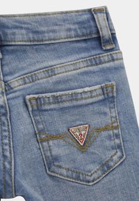 Ljust blå denimjeans med en triangulär röd och vit "GUESS"-logotypapplikation på bakfickan, med kontrasterande gula söm detaljer.