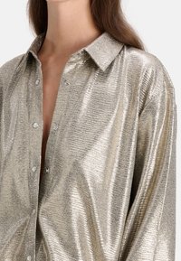 Glänzende, metallische silberne Button-Up-Hemd mit einem scharfen Kragen und einer lockeren Passform, das über tonale Streifen und perlenartige Knöpfe verfügt.