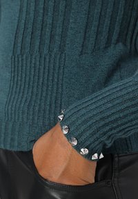 Pull côtelé bleu sarcelle avec des clous pyramides métalliques au niveau du poignet ; texture douce, forme ajustée et couture détaillée. Gros plan sur la manche et la main.