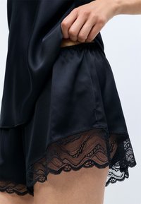 Pijamas de satén negro con un top y pantalones cortos de corte relajado, adornados con encaje ondulado en el dobladillo. Cintura elástica para mayor comodidad.