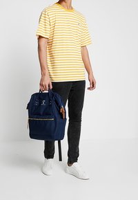 Námořnický batoh se zipem a béžovými koženými akcenty, nesený osobou v pruhovaném žlutém a bílém tričku a černých džínách.