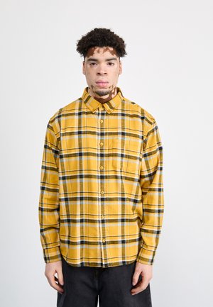 Camisa de franela a cuadros en mostaza con acentos en negro y azul, de mangas largas, con botón en la parte delantera y un solo bolsillo en el pecho.