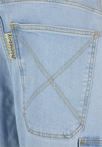Lyseblå denim jeans med en baklomme med gul søm og en flerfarget etikett. Teksturen er glatt med et subtilt vevemønster.
