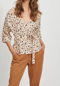 Blouse beige clair avec un design croisé, dotée d'une ceinture à nouer, de manches 3/4 et d'un motif abstrait coloré. Associée à un pantalon marron.
