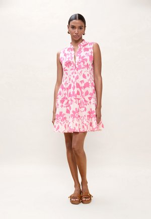 Mujer de pie, vistiendo un vestido sin mangas de flores rosas y blancas, y sandalias marrones con plataforma, sobre un fondo claro y liso.