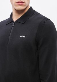 Črna polo majica na zadrgo iz teksturirane tkanine, z ostrim ovratnikom in belim napisom "BOSS" na levi strani prsnega koša.