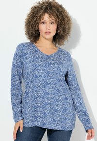 Una donna con i capelli ricci indossa una maglietta a maniche lunghe blu con motivi floreali e jeans scuri, in piedi di fronte a uno sfondo bianco uniforme.
