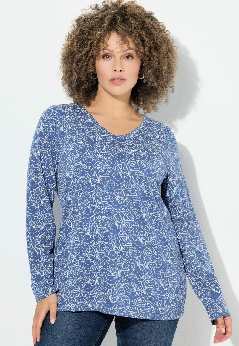 Una donna con i capelli ricci indossa una maglietta a maniche lunghe blu con motivi floreali e jeans scuri, in piedi di fronte a uno sfondo bianco uniforme.