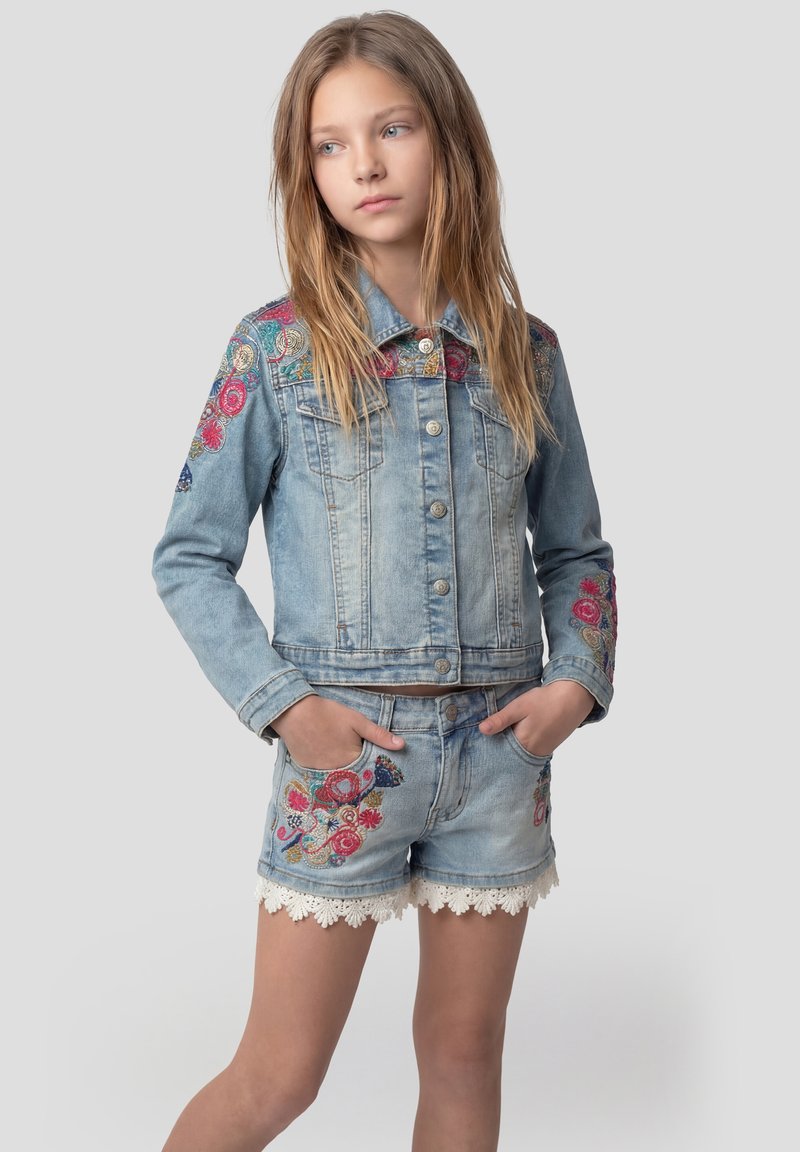 Ragazza giovane che indossa una giacca di jeans azzurro chiaro e pantaloncini con ricami floreali colorati e bordi in pizzo bianco, con le mani in tasca, che guarda di lato.