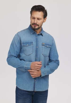 Mężczyzna w jasnoniebieskiej dżinsowej koszuli zapiętej na guziki i ciemnoniebieskich dżinsach, poprawiający lewy mankiet prawą ręką.