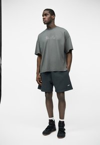 Jeune homme debout, mains dans les poches, portant un t-shirt gris surdimensionné, un short foncé, des chaussettes blanches et des baskets montantes noires.