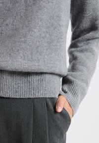 Szary dziany sweter z prążkowanymi mankietami i dołem, wykonany z miękkiego materiału, noszony na ciemnoszarych spodniach z ręką spoczywającą w kieszeni.